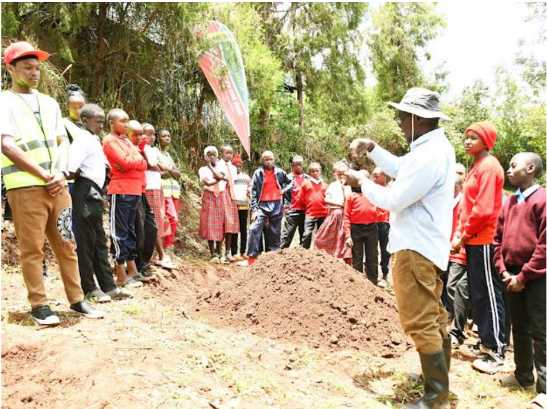 A man in a sun hat teaching schoolchildren gathered around a mound of rich soil in a wooded rural setting