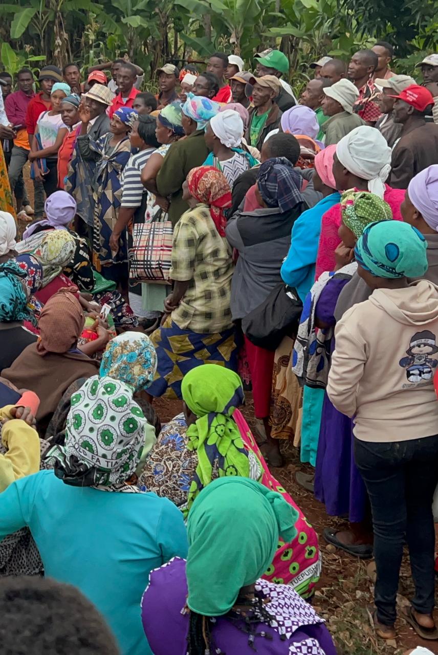 A large community gathering of women and men in rural Kenya, listening under banana trees